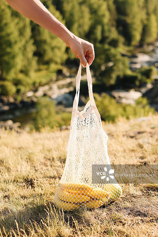 一只手拿着可重复使用的网状袋，装着水果，背景是绿色森林，户外环保图片素材