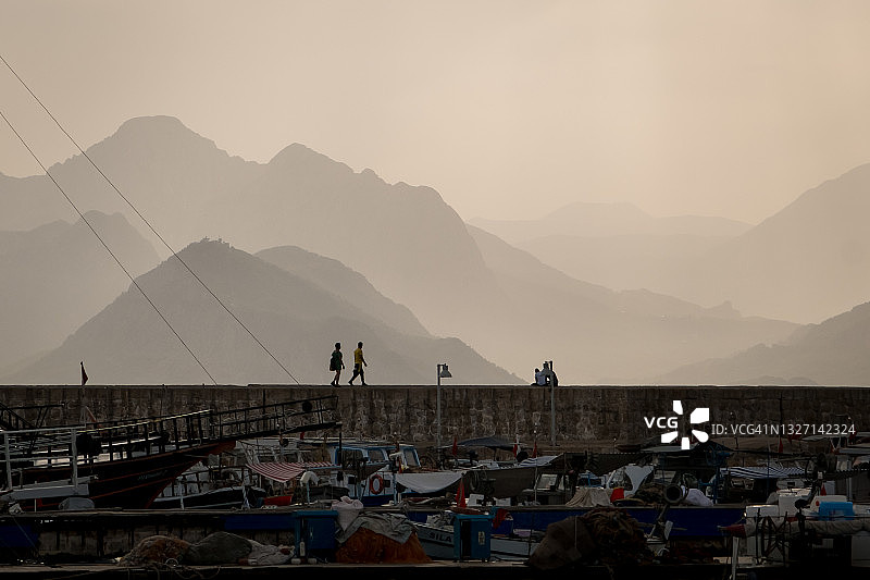 地平线上层叠的山脉，阳光穿透云层。日落前的傍晚，土耳其安塔利亚码头上的人影图片素材