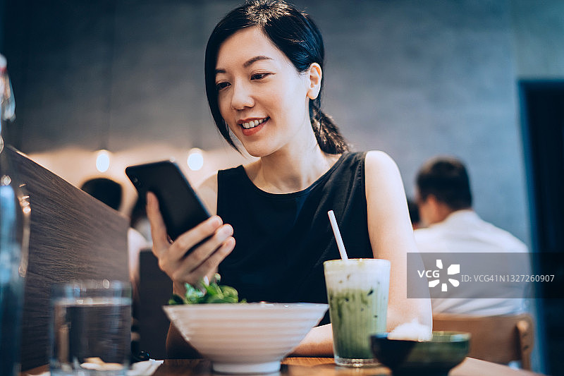 年轻亚洲女人在餐厅使用智能手机，享用新鲜的纯素沙拉和绿色冰沙，享受健康膳食。绿色生活方式。素食概念图片素材
