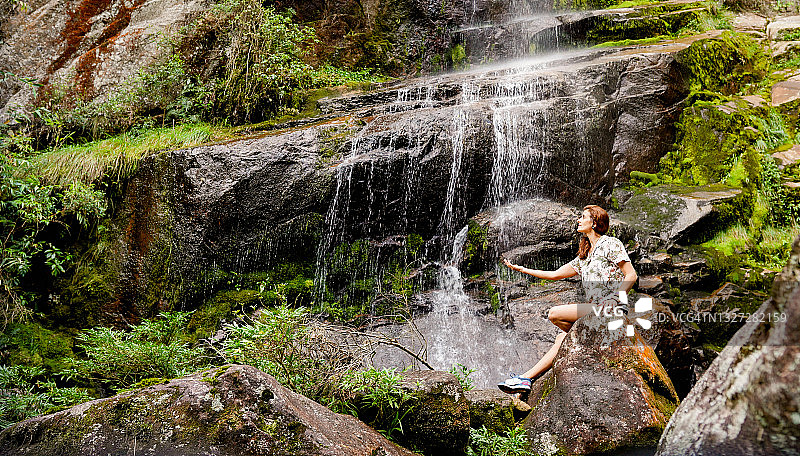 女人坐在岩石上，伸手触摸瀑布图片素材