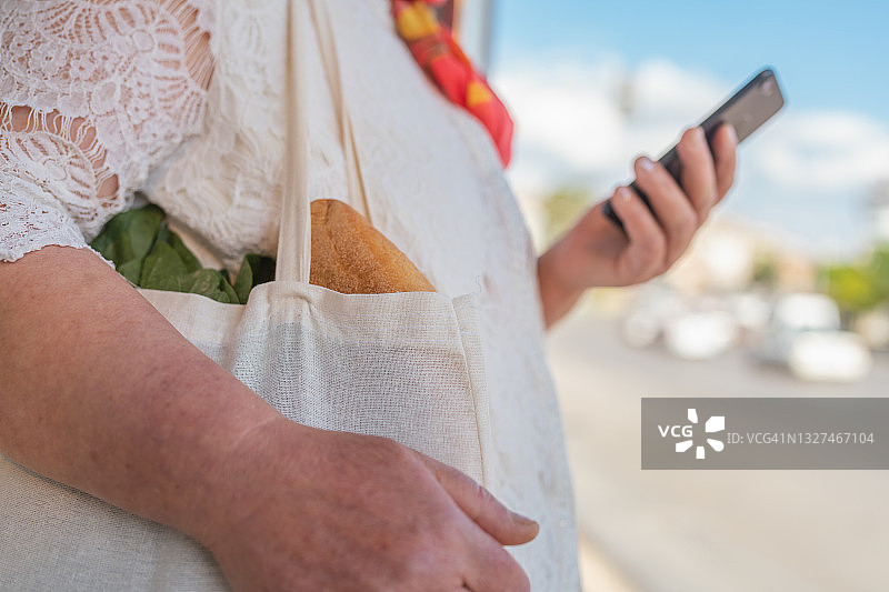 在公交车站等车时，携带可重复使用购物袋并使用智能手机的穆斯林妇女图片素材