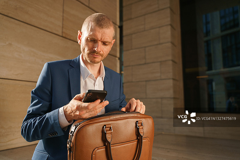 身穿蓝色西装的年轻英俊商务男人打开棕色笔记本电脑包并查看手机的特写图片素材