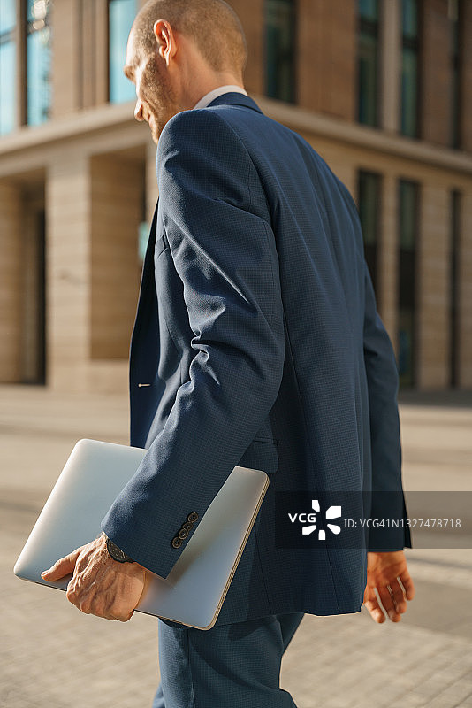 一位穿着蓝色西装、手提笔记本电脑的年轻商务男士走在商业中心附近的街道上的背影图片素材