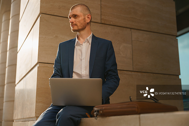 身穿蓝色西装的年轻经理坐在圆柱旁，看向远方图片素材