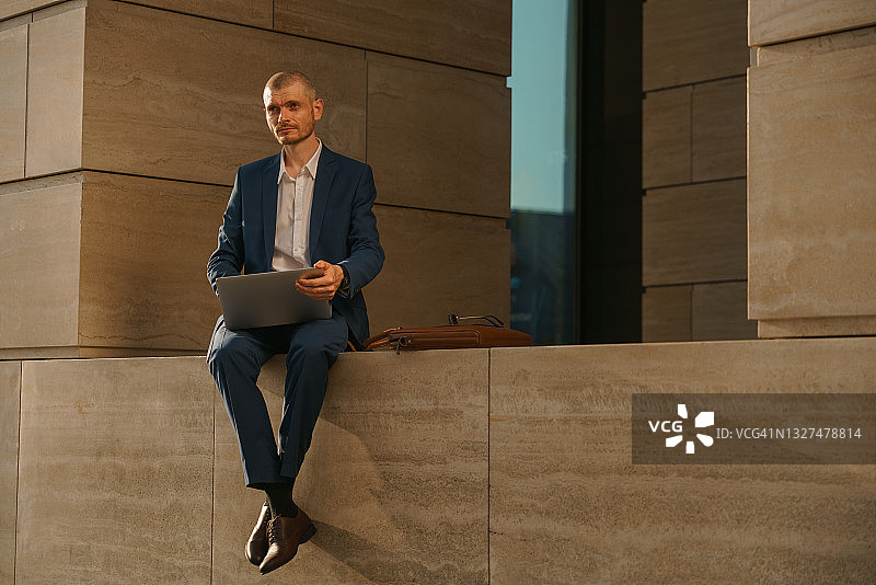 有魅力的苗条办公室经理坐在商务中心的外墙上，带着笔记本电脑看着镜头图片素材