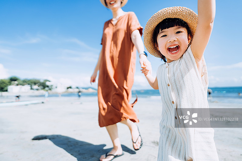 充满爱意的年轻亚裔母亲和快乐的小女儿手牵手走在阳光明媚的夏日海滩上，分享美好时光图片素材