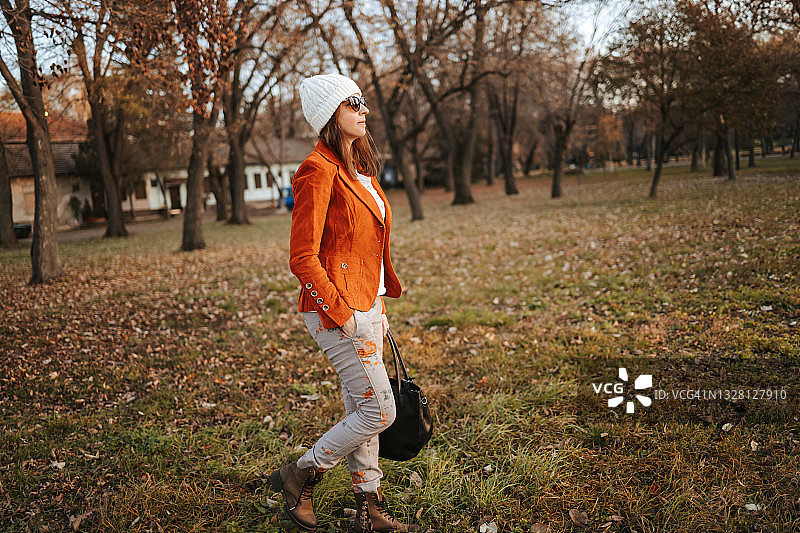 身穿橙色夹克的时尚年轻女子在秋季公园里散步，眺望远方图片素材