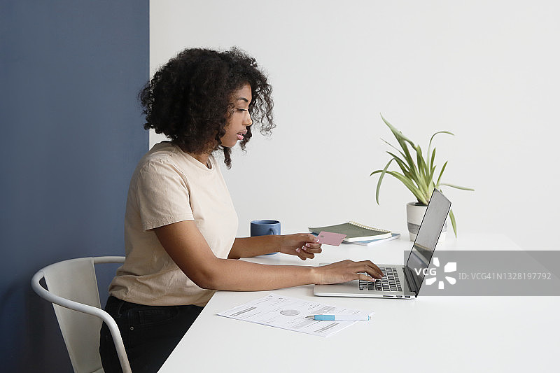 一位有卷发的非裔美国千禧一代女性在家里使用信用卡在笔记本电脑上付款，身着休闲服装图片素材