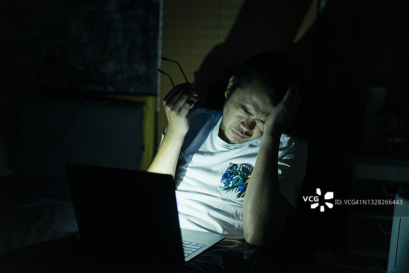 在卧室里使用笔记本电脑，感到疲惫不堪、工作过度的亚洲青年图片素材