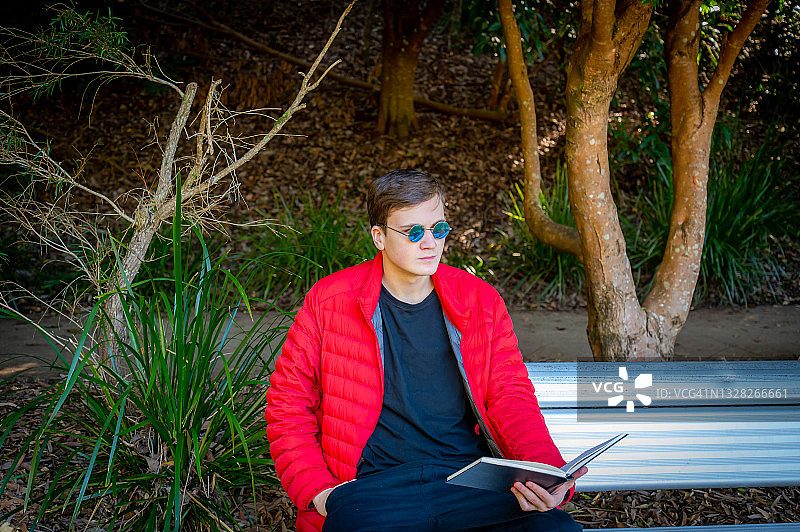 穿红色羽绒服戴墨镜的少年在公园里阅读图片素材