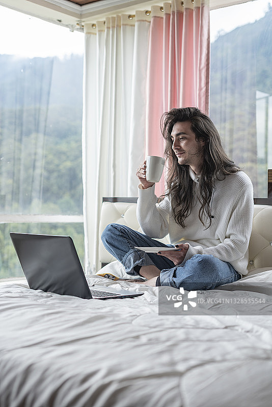 坐在床上用笔记本电脑学习，喝咖啡的拉丁裔男人图片素材