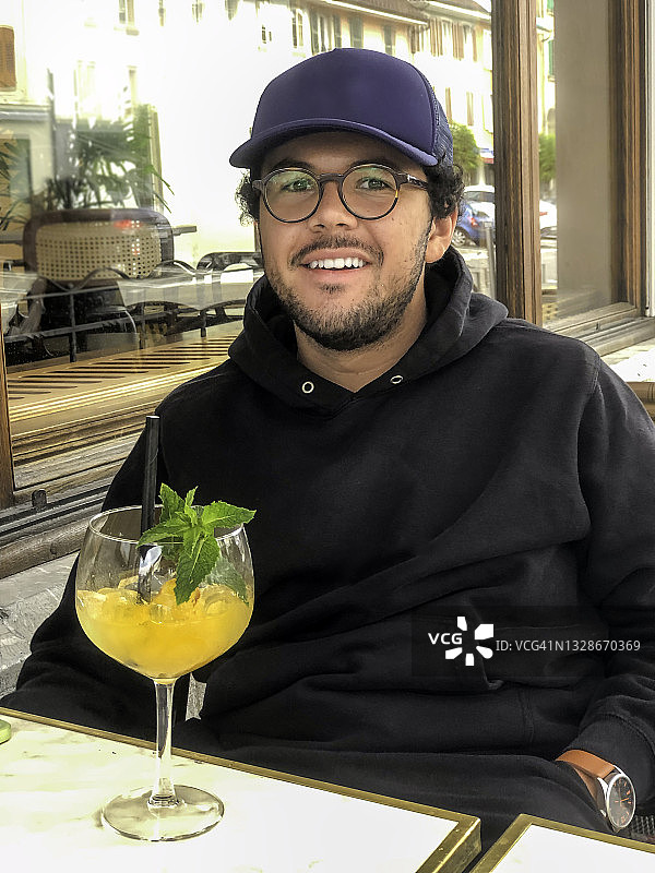 一位戴着胡须、眼镜、蓝色帽子和黑色帽衫的18岁男学生在户外咖啡馆露台上喝水果鸡尾酒的肖像图片素材