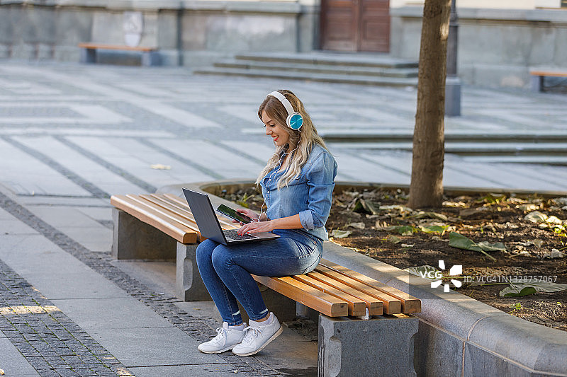 戴着耳机在公园使用笔记本电脑进行远程工作的年轻女人图片素材