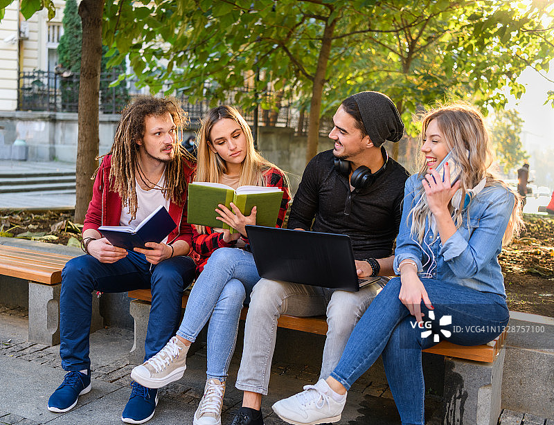 美丽的年轻女人们和她们的男友坐在公园的长椅上用笔记本电脑工作。图片素材