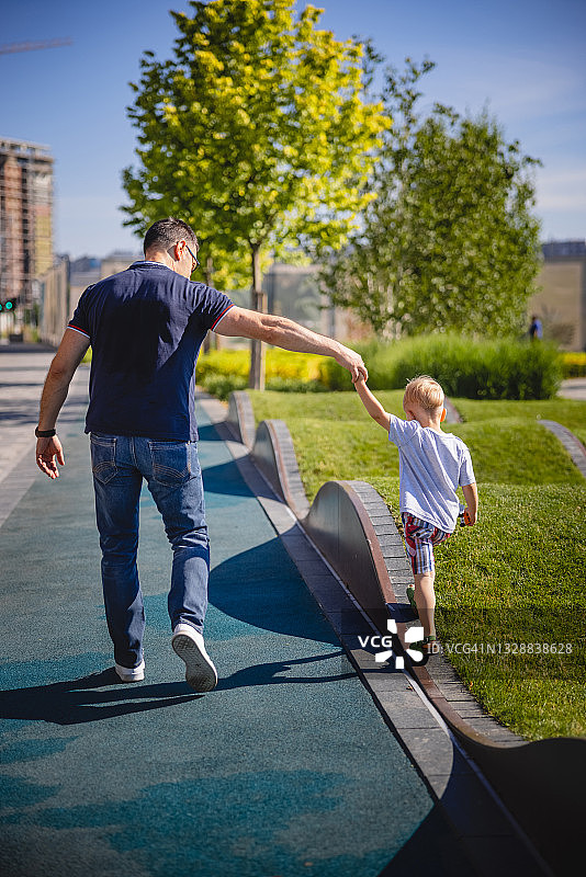 父亲在公园里和蹒跚学步的孩子玩耍，牵着他的手帮助他爬墙的背影图片素材