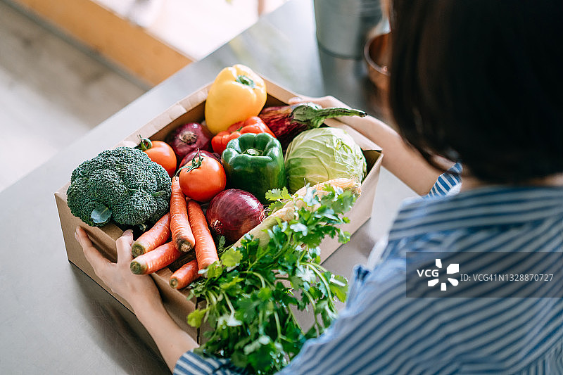 年轻亚洲女性在厨房拿着装满彩色新鲜有机食品盒子的肩上视角图片素材