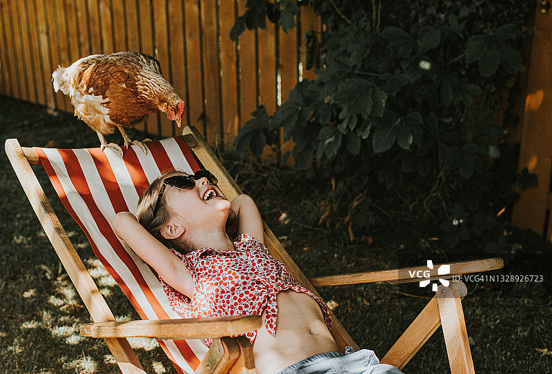 放松的孩子躺在躺椅上，鸡站在躺椅边缘看着她的脸，她笑着图片素材