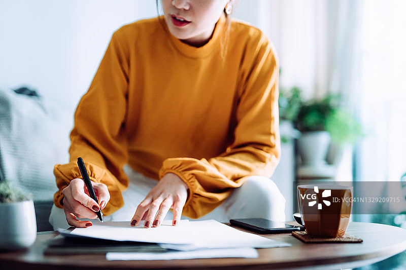 年轻亚洲女性在家中客厅拿着笔签署文件的局部照片，交易概念图片素材