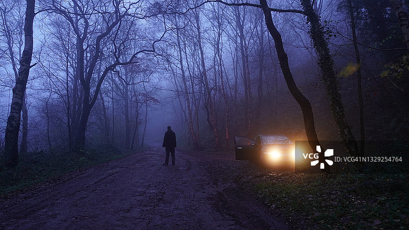 神秘剧情概念：男子在雾冬之夜，森林中停放的汽车旁凝视着幽深的小路图片素材