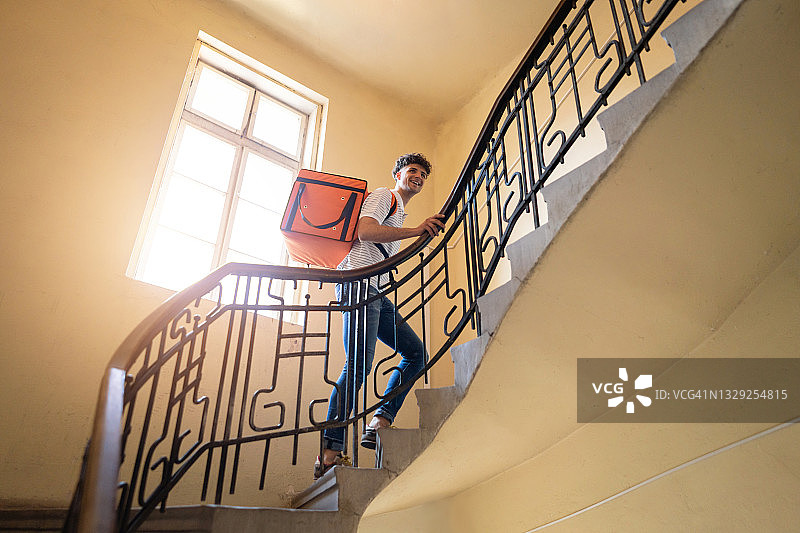 一个白人快递员提着红色的包在住宅楼的楼梯上行走图片素材