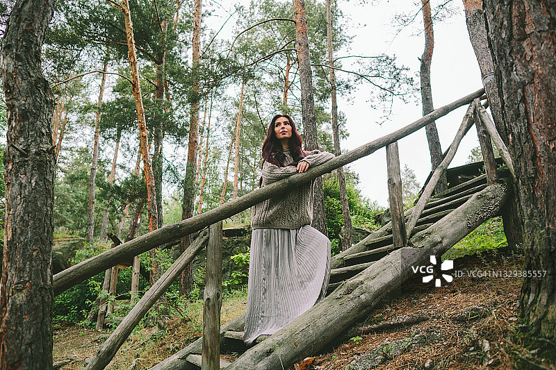 站在森林木梯上的银色长裙棕发年轻女子图片素材