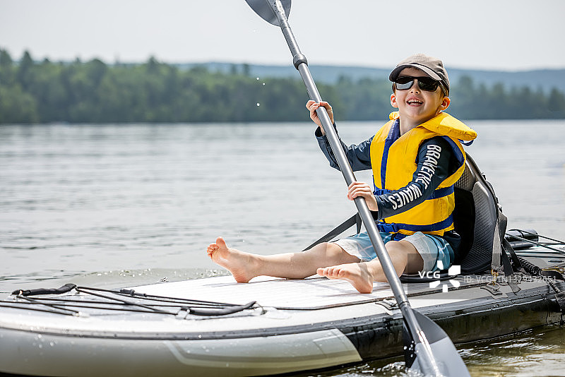 在温暖阳光明媚的夏日度假时，在桨板上划皮划艇的红发小男孩图片素材