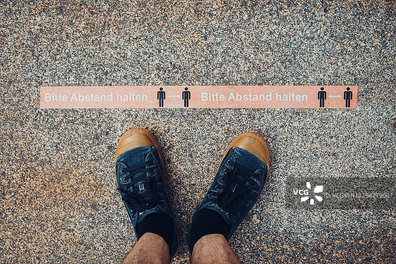 男子穿着蓝色运动鞋站在用德语写的安全距离标志前，由于新冠疫情爆发，为了保持安全的社交距离图片素材