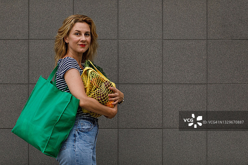 拿着两个装满杂货的可重复使用袋子的女人，对着镜头微笑图片素材
