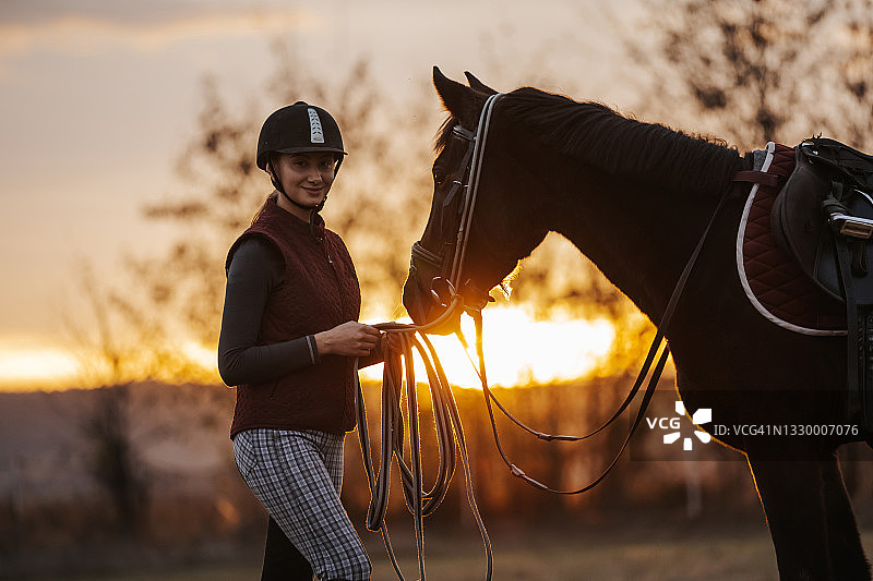 年轻女骑师在牧场上牵着赛马，背景是美丽的日落图片素材