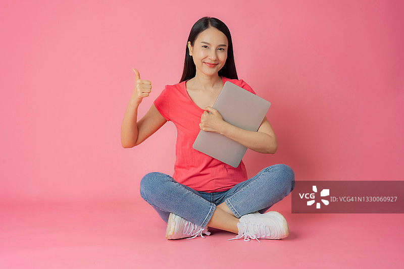身穿红衣牛仔裤的亚洲女性坐在粉色背景上，手持笔记本电脑图片素材