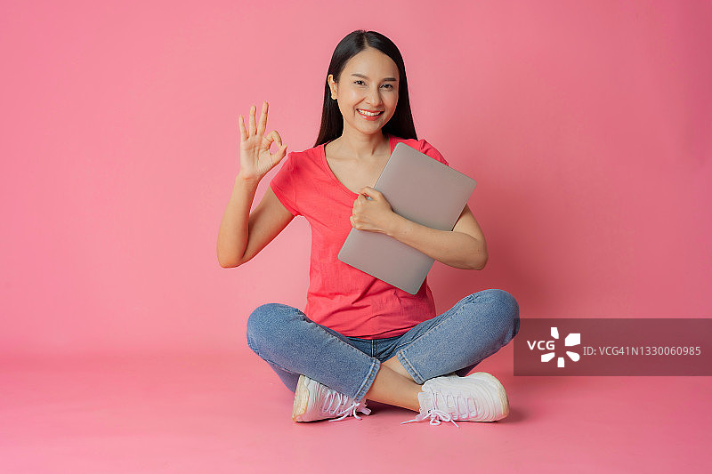 身穿红衣牛仔裤的亚洲女性坐在粉色背景上，手持笔记本电脑图片素材