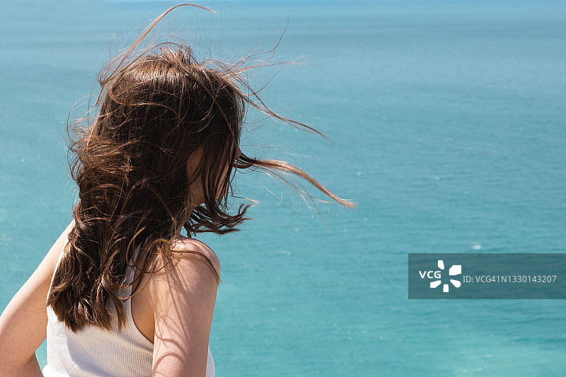 年轻漂亮的女人身穿白色T恤和灰色牛仔裤坐在悬崖边欣赏海景，后视图图片素材