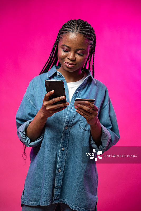 深肤色女孩在粉色摄影棚内使用手机信用卡在线购物。图片素材