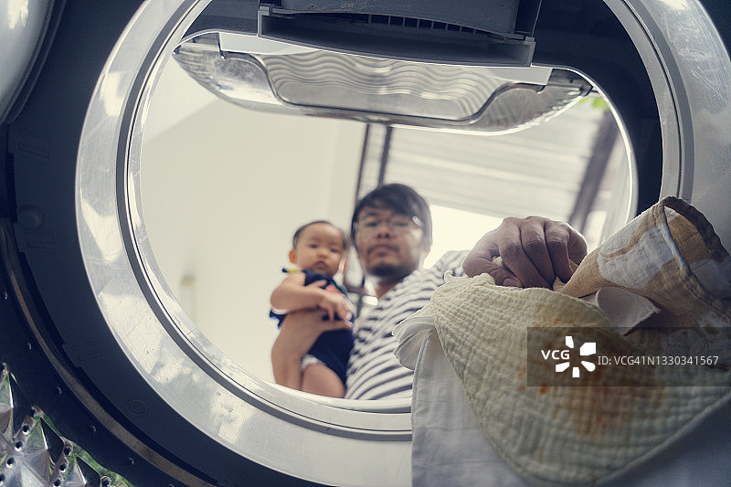 洗衣机内部的低角度视角：亚裔单身父亲拥抱女儿，对婴儿围裙的脏污和频繁清洁感到担忧和厌烦图片素材