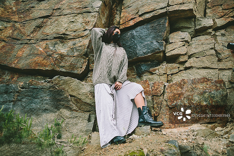 一位留着长发的年轻棕发女子，身穿银色长裙和毛衣，在石坡背景前戴着黑色医用口罩对着镜头摆姿势。疫情期间的蒙面旅行。图片素材