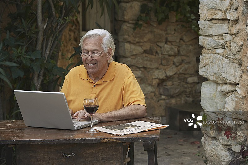 年长的男人在露台的桌旁一边喝红酒一边用笔记本电脑图片素材