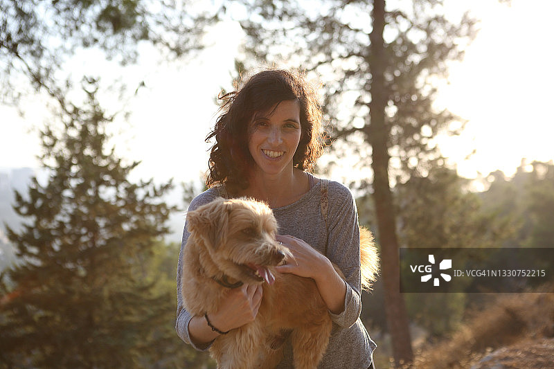 在松树林中抱着梗犬，在日落时幸福微笑的年轻女人图片素材