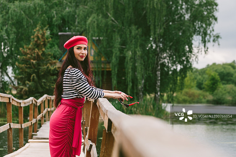 年轻女子戴红色贝雷帽，在湖边木桥上手持复古手提箱眺望远方图片素材