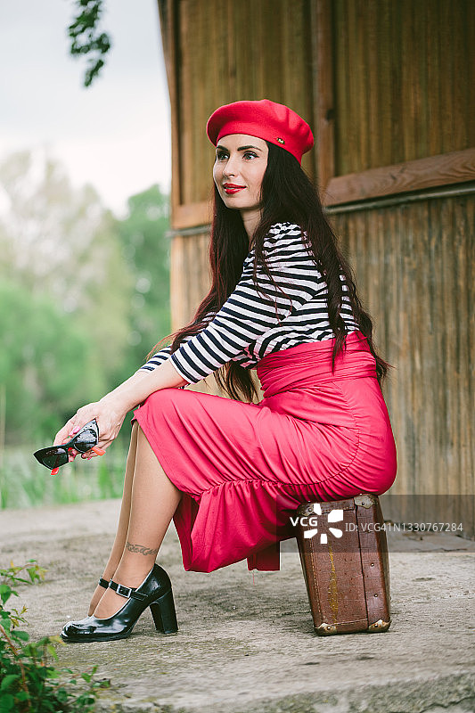 棕发女子戴红色贝雷帽和黑框眼镜，身穿条纹外套、红色裙子和黑色鞋子，坐在木屋旁的复古手提箱上休息图片素材