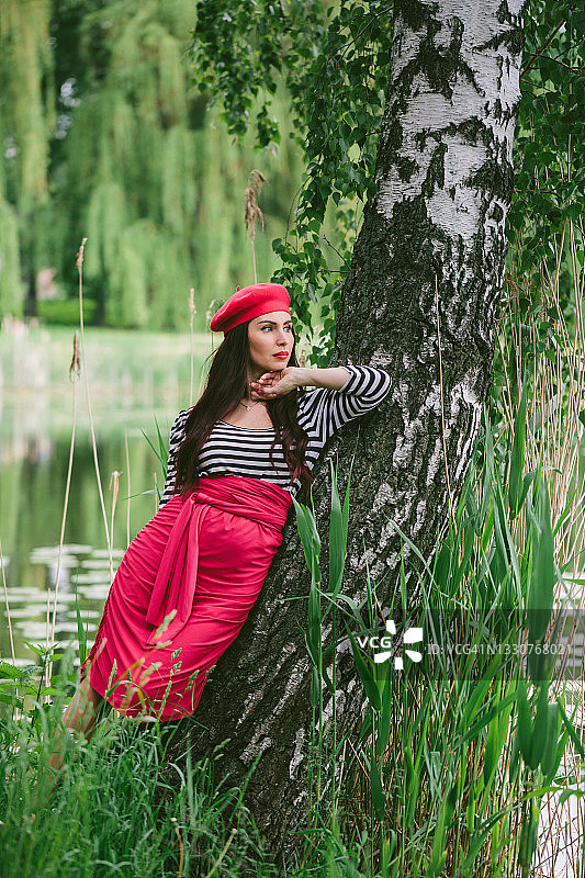 一位年轻的黑发女子，头戴红色贝雷帽，戴着黑色眼镜，穿着条纹夹克和红色裙子，靠在湖边绿色森林中的桦树上，手托着下巴，向后看图片素材