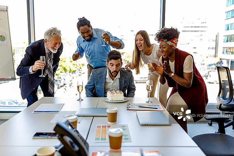 男人在办公室和同事一起庆祝生日，并吹灭蛋糕上的蜡烛图片素材