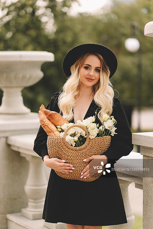 一位美丽可爱的年轻女子，金色卷发，化着精致的妆容，戴着帽子，穿着优雅的黑色连衣裙，拎着装满鲜花和法棍面包的柳条包，在城市剧院和建筑附近的植物和树木中漫步图片素材