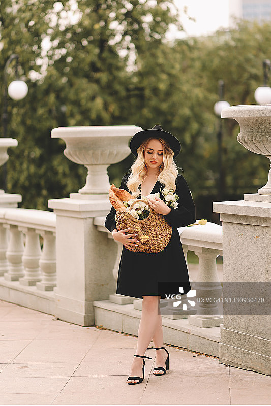 一位美丽可爱的年轻女子，金色卷发，化着精致的妆容，戴着帽子，穿着优雅的黑色连衣裙，拎着装满鲜花和法棍面包的柳条包，在城市剧院和建筑附近的植物和树木中漫步图片素材