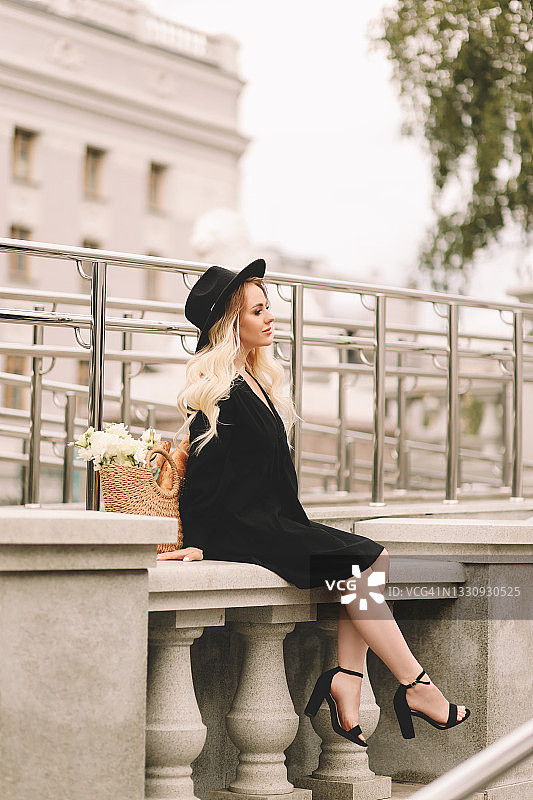 一位美丽可爱的年轻女子，金色卷发，化着精致的妆容，戴着帽子，穿着优雅的黑色连衣裙，拎着装满鲜花和法棍面包的柳条包，在城市剧院和建筑附近的植物和树木中漫步图片素材