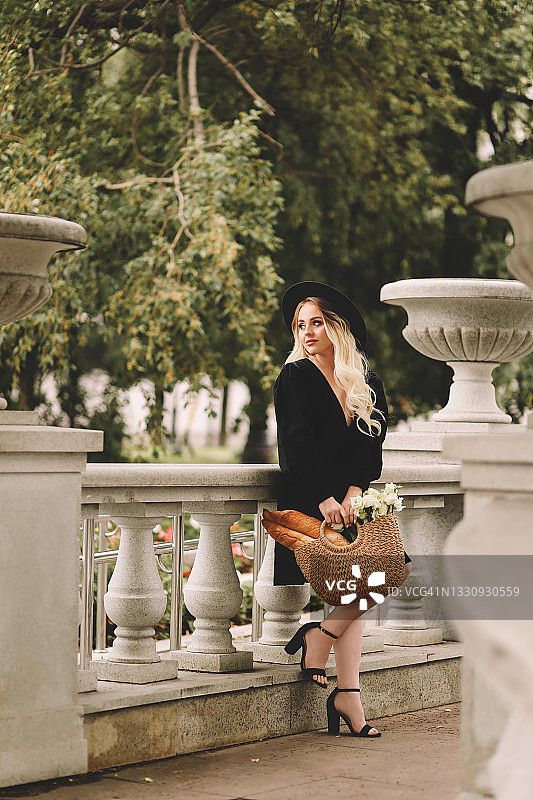 一位美丽可爱的年轻女子，金色卷发，化着精致的妆容，戴着帽子，穿着优雅的黑色连衣裙，拎着装满鲜花和法棍面包的柳条包，在城市剧院和建筑附近的植物和树木中漫步图片素材