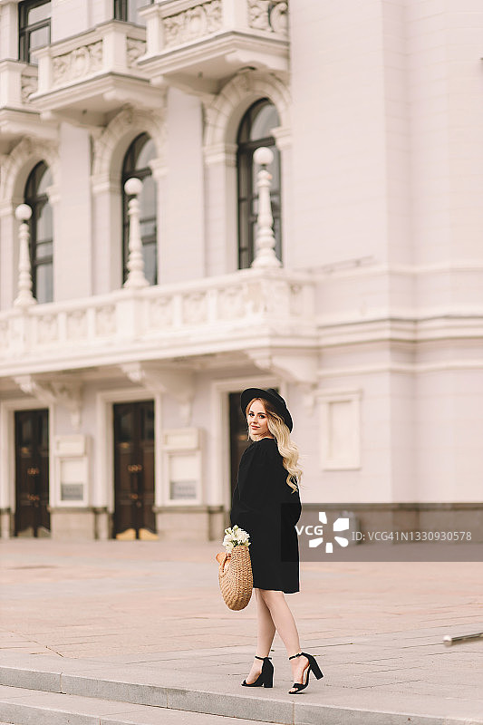 一位美丽可爱的年轻女子，金色卷发，化着精致的妆容，戴着帽子，穿着优雅的黑色连衣裙，拎着装满鲜花和法棍面包的柳条包，在城市剧院和建筑附近的植物和树木中漫步图片素材