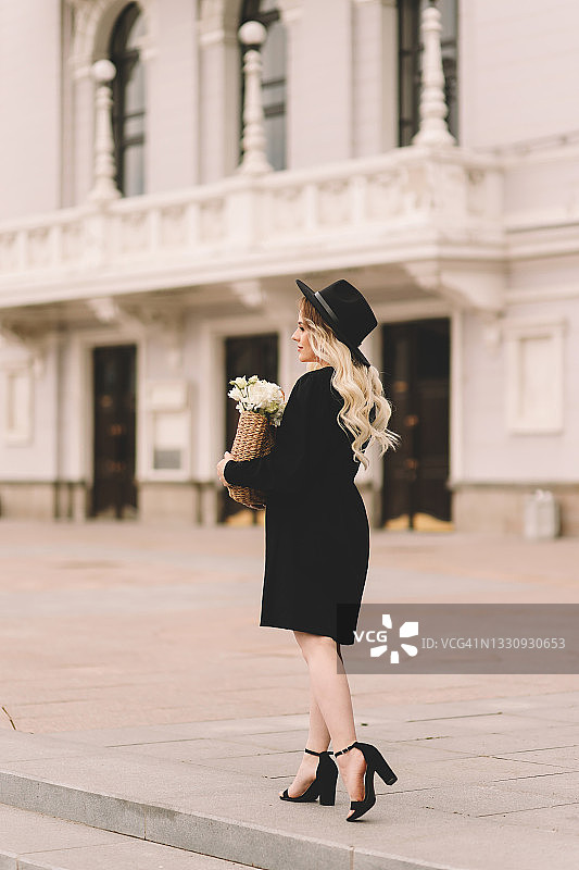 一位美丽可爱的年轻女子，金色卷发，化着精致的妆容，戴着帽子，穿着优雅的黑色连衣裙，拎着装满鲜花和法棍面包的柳条包，在城市剧院和建筑附近的植物和树木中漫步图片素材