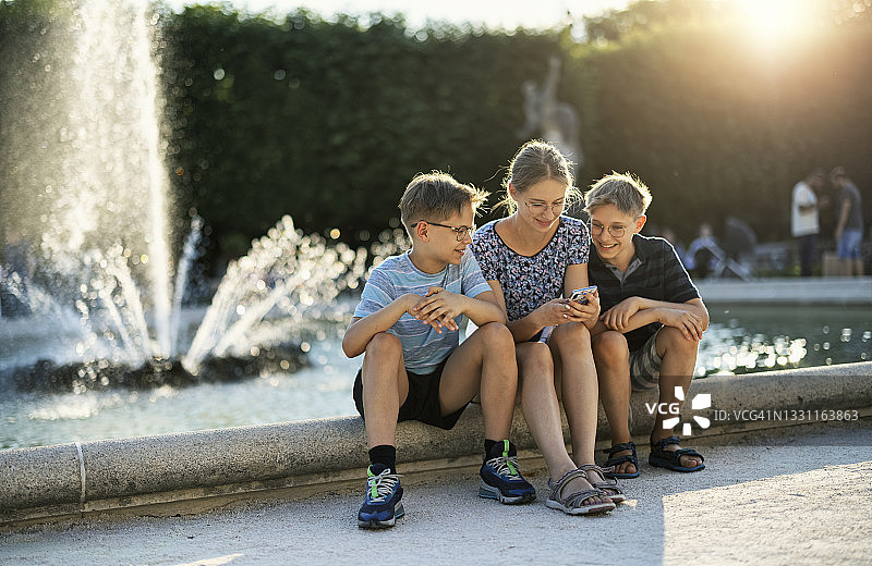 三个孩子在公园喷泉附近查看智能手机图片素材