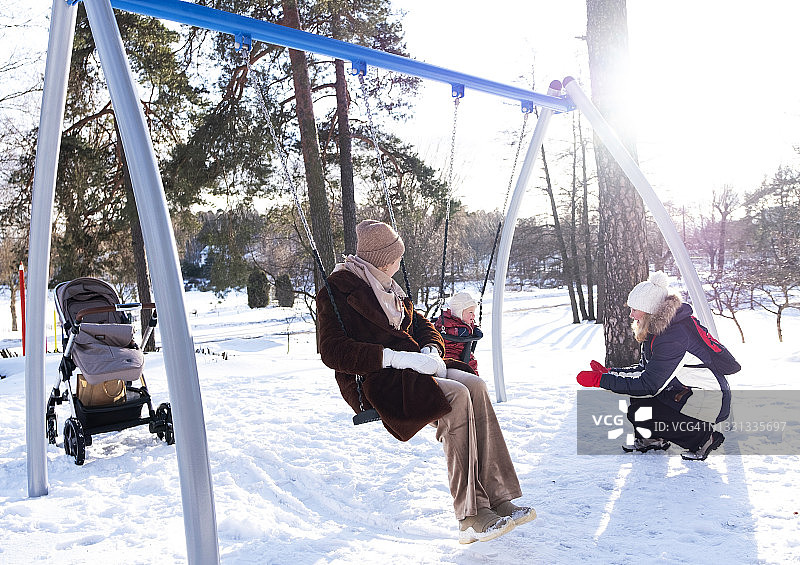 两位年轻女子和一位婴儿在操场上，女子在秋千上摇晃婴儿。图片素材