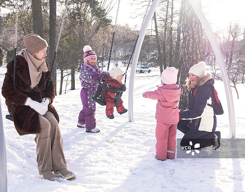 冬日游乐场：两家人带孩子散步，白人小女孩在荡秋千，父母在一旁看护图片素材